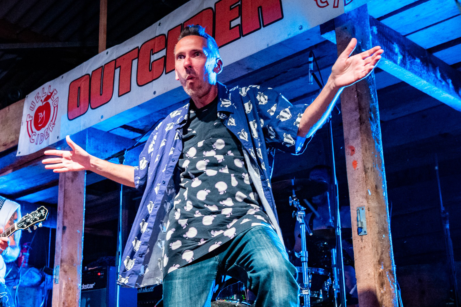 Kev Davis from the band Skimmity Hitchers on stage with his arms outstretched and hands open while wearing blue jeans and a blue and and white t-shirt with another open blue and white button-up shirt over the top 